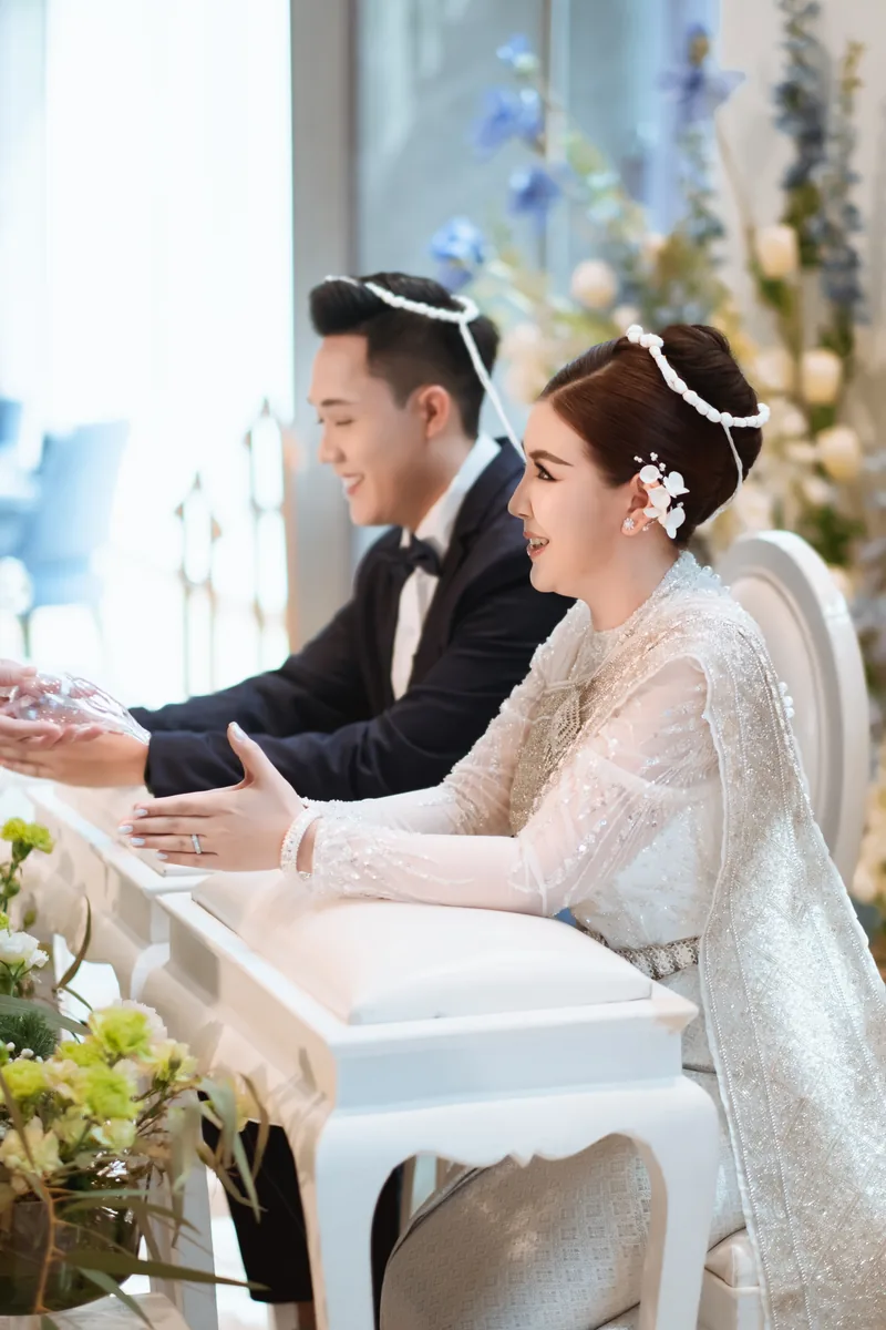 บ่าวสาวในชุดไทยระหว่างพิธีรดน้ำสังข์ ฉากหลังเป็นซุ้มดอกไม้สีขาวฟ้า